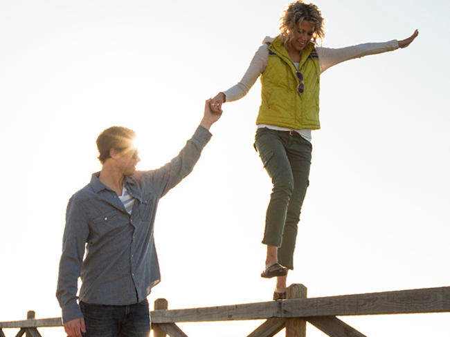 A woman walks on top of a wooden fence, her husband holding hand to help her balance